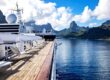 Cruise ship sun deck with loungers overlooking a calm tropical lagoon and dramatic green mountains under a blue sky.