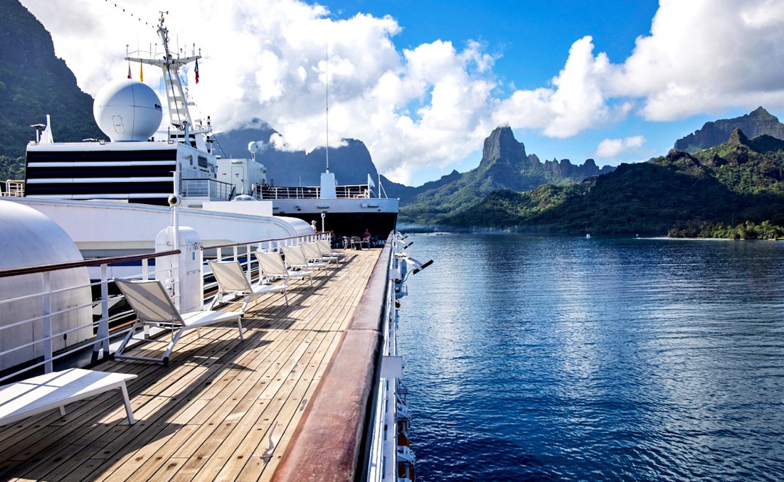 Cruise ship sun deck with loungers overlooking a calm tropical lagoon and dramatic green mountains under a blue sky.