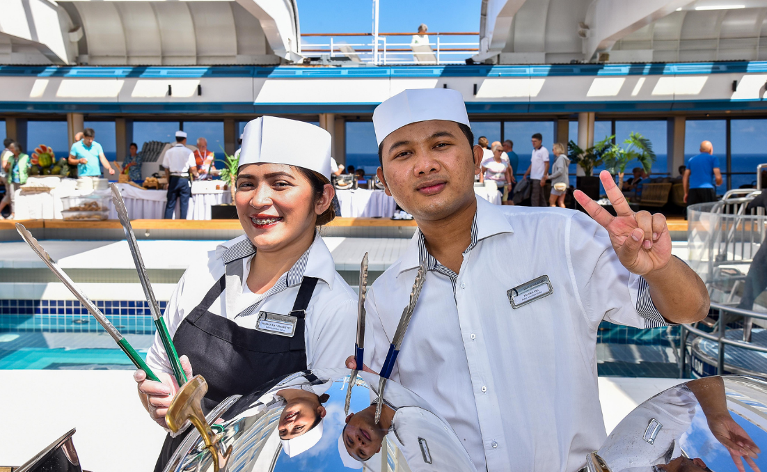 Zwei Crewmitglieder aus Hospitality/Galley in Uniform lächeln am Pooldeck, im Hintergrund ein Service-Setup und Blick aufs Meer.