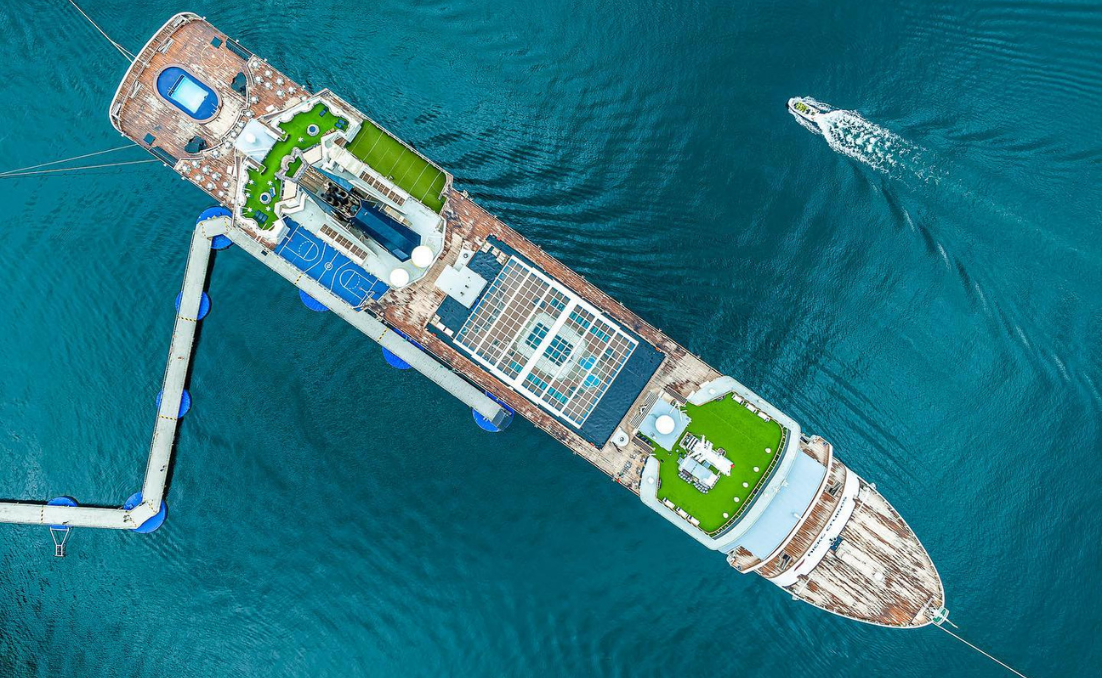 Aerial view of the Vasco da Gama ship alongside a pier, with the deck visible over blue water and a small motorboat in the distance.